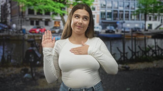 Woman raising right hand and placing left hand on chest on a city street by a canal with parked bikes and buildings; oath sincerity.