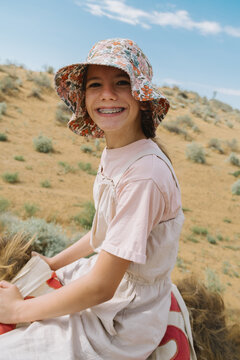A smiling girl wearing a floral hat riding a camel in a desert.