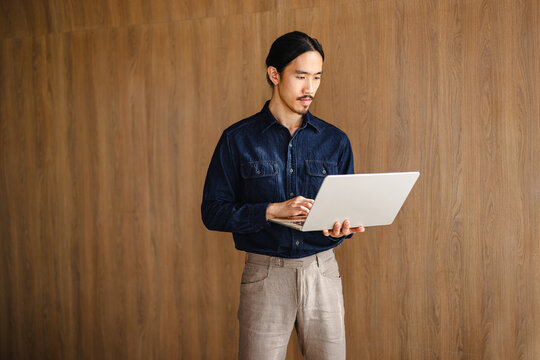 Man Using Laptop While Standing in Indoor Setting With Wooden Wall