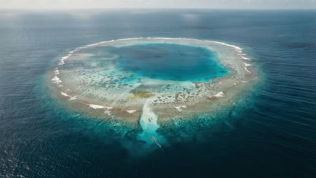 Aerial view of a circular coral reef atoll surrounded by deep blue ocean water, featuring a narrow channel leading into the turquoise lagoon