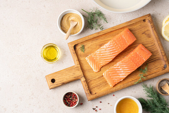 Fresh raw salmon fillet on wooden cutting board surrounded by ingredients for honey mustard lemon marinade, including lemon, mustard, honey and herbs. High protein healthy food