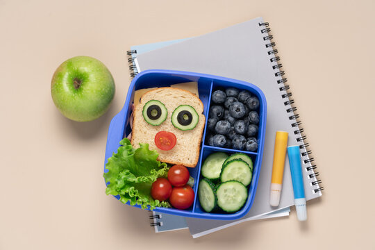 Kids school lunch box with fun face sandwich vegetables and blueberries in blue container on light beige background, back to school concept