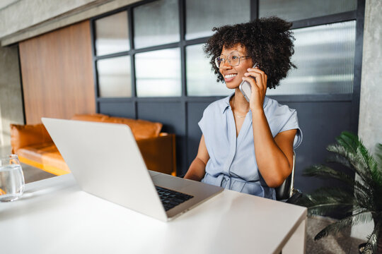 Woman Talks on Phone While Sitting at Desk With Laptop in Office