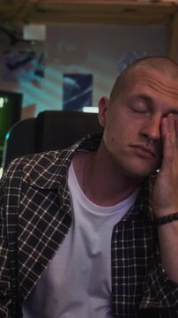 Vertical video of exhausted young adult Caucasian man taking off headphones and glasses, rubbing eyes after late night gaming. Ideal for campaigns on screen addiction and digital burnout