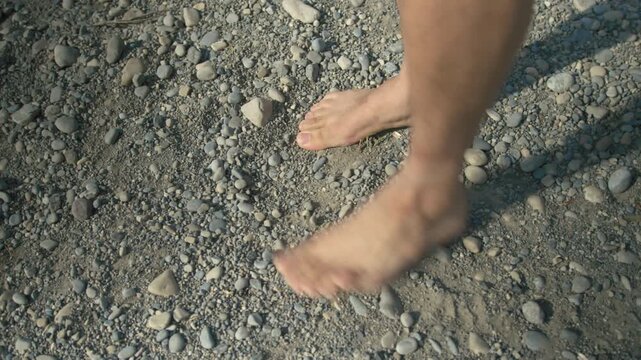 Close-up of a man walking barefoot on a pebble riverbank in warm golden light