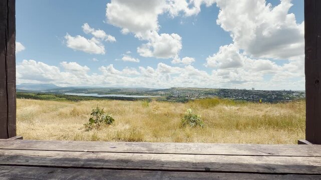 A wooden frame overlooks a grassy hill and lake in Crimea. White clouds drift across the bright blue sky. This wide open view fills the heart with peaceful joy and freedom.