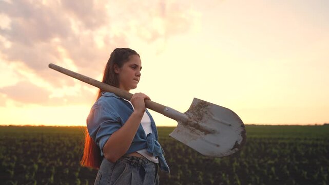 A teenage farmer girl stands in a field at sunset, holding a shovel as she gazes thoughtfully across the farmland. The teenage farmer girl represents hard work and dedication.