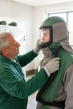 Experienced supervisor helping worker put on protective gear for sandblasting. Professional industrial safety assistance in workshop. Protective equipment suit and helmet for abrasive job.
