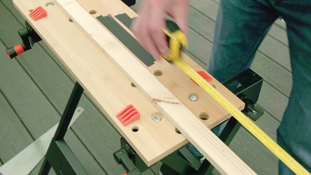 Woodworker measures lumber on a workbench. Hands grip tape measure, pencil ready. Green deck background, outdoor workshop. Scene conveys precision in DIY projects. No animals or plants present. 1-6