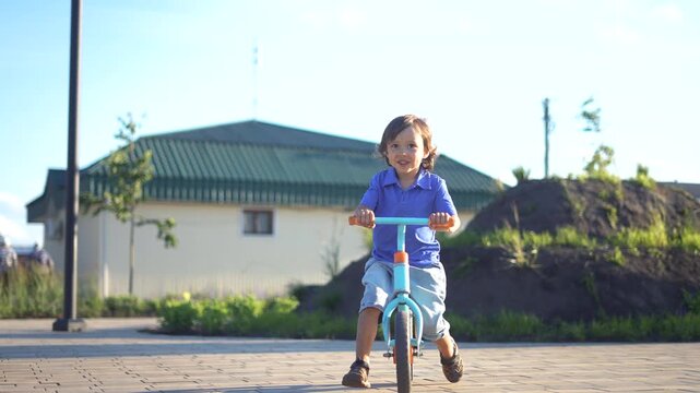 A boy rides a blue balance bike in a sunny setting, enjoying the outdoors. The boy smiles as he navigates a paved path near a green-roofed house.