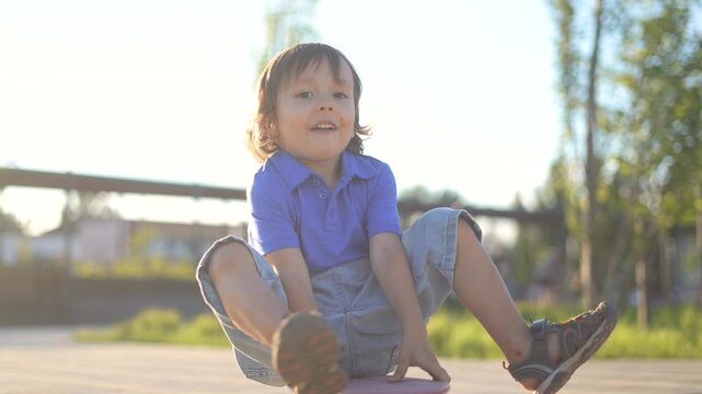 The boy squats on the skateboard, smiling as he balances in the sunlight. The boy enjoys the outdoors, while greenery surrounds the playful scene.