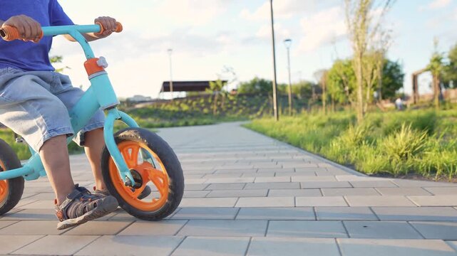 A boy rides a blue bike with orange wheels. The boy enjoys riding the bike on a paved path surrounded by grass and trees.