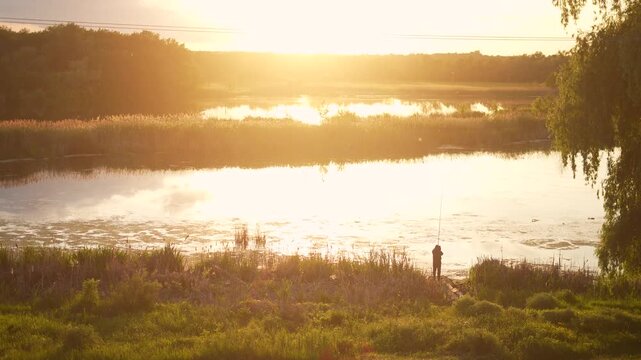 A man stands by the water, casting a fishing rod into the calm water. The man enjoys fishing as the sun sets, illuminating the tranquil water.