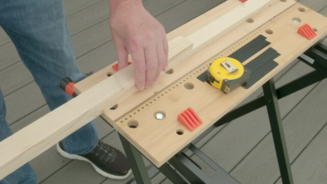 Man adjusts wood on workbench with tape measure. Hand guides plank over marked surface. Tool setup shows precise measurement process. DIY project scene for carpentry or home improvement. Two of six in