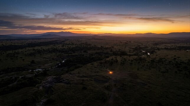 Aerial view of a remote African savanna at sunset with mountain silhouettes, winding stream, scattered trees, and a small campfire creating a wilderness travel concept