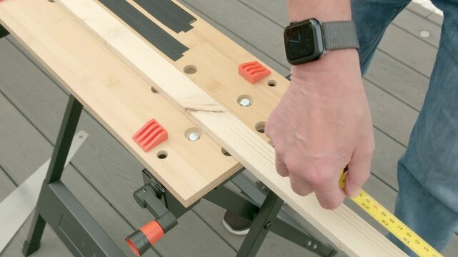 Person measures wood on a workbench. Hands hold ruler, aligning it with the wood. Workbench holds tools and clamps securely. Outdoor setting suggests DIY or home improvement projects. Five of six 