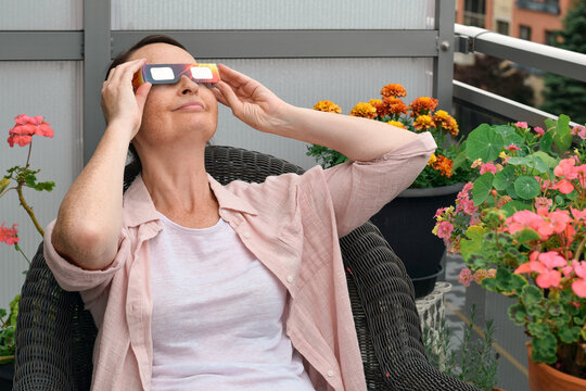 Middle age woman sitting in chair on balcony wearing solar eclipse glass, smiling and looking up at sky surrounded by potted flowers