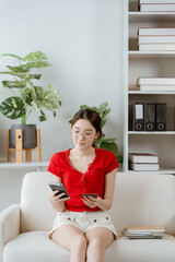 Young Woman Managing Finances: A young woman with glasses, wearing a red top and white shorts, sits on a couch and calculates her finances using a calculator and credit card. © Wicitr