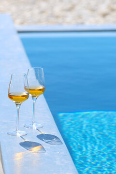 Two glasses of Greek orange wine rest on a sunlit poolside ledge, their reflections shimmering against bright blue water in a tranquil summer scene of leisure and relaxation.