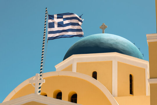 A Greek flag waves proudly beside a yellow church with a blue dome and cross under a clear sky, symbolizing national pride and island architecture.