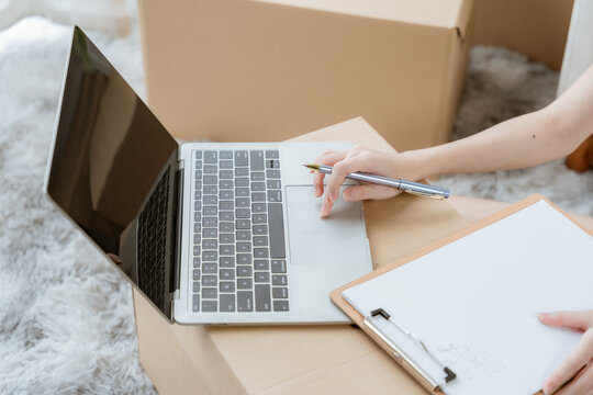 Unpacking and Organizing: A close-up shot of hands writing on a clipboard next to an open laptop, surrounded by cardboard boxes, suggesting moving or organizing items.