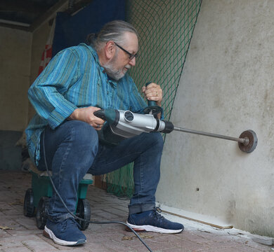 An older man, wearing glasses and a blue striped shirt, is sitting on a wheeled cart while using a power drill to bore into a brick wall. 
