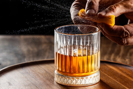 Hands pressing an orange peel releasing the oils on top of an old fashioned cocktail on a wooden bar top.