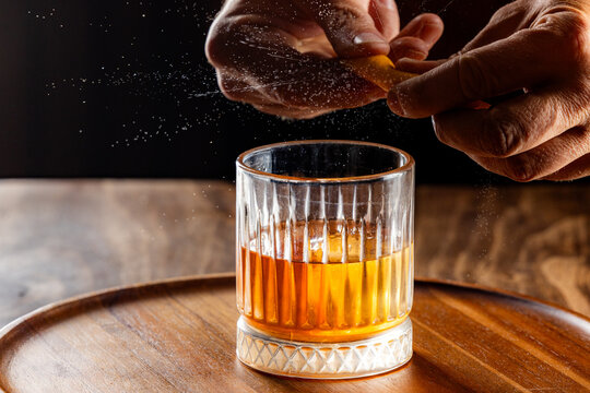 Hands pressing an orange peel releasing the oils on top of an old fashioned cocktail on a wooden bar top.