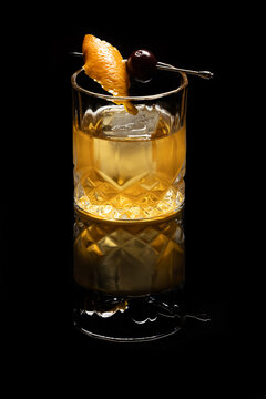 Close up of a classic old fashioned cocktail on a dark reflective background garnished with a cherry and an orange twist.