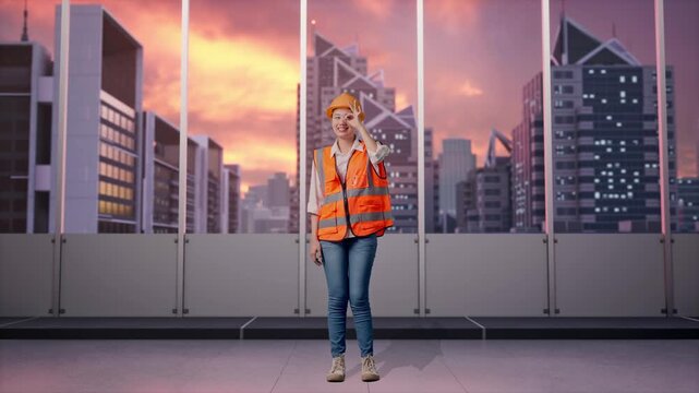 Full Body Of Asian Female Engineer With Safety Helmet Showing Ok Hand Sign Over Eye And Smiling To Camera While Standing at Office with Panoramic City Skyline at Sunset