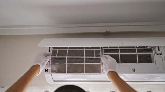 Skilled technician's hands in white gloves carefully removing a very dusty and dirty filter from a wall-mounted air conditioner unit during routine hvac system maintenance