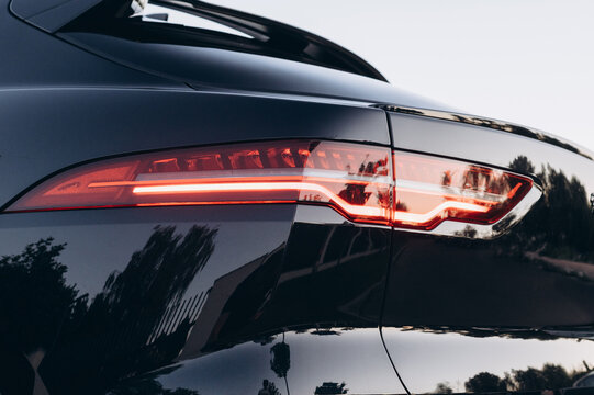 Close up of a black car rear with an illuminated red taillight, sleek bodywork, reflective surface, and rear spoiler in soft evening light.