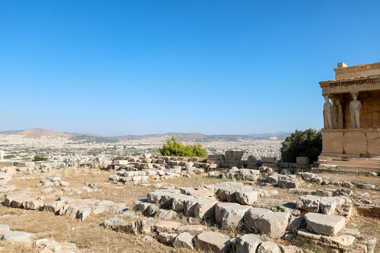 Ancient stone ruins on the Acropolis of Athens stretch toward the horizon with the Erechtheion temple visible at right, overlooking the modern city under a bright blue sky.
