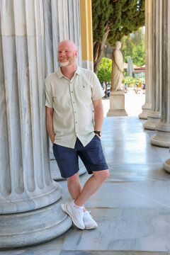 A man in casual summer clothing leans against a marble column in a neoclassical walkway, smiling in bright sunlight surrounded by historic architecture in Athens.