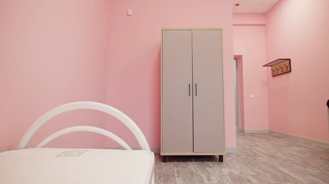 Metal bed with mattress rests near tall wooden wardrobe cabinet. Open doorway reveals bright adjoining room with pastel pink walls