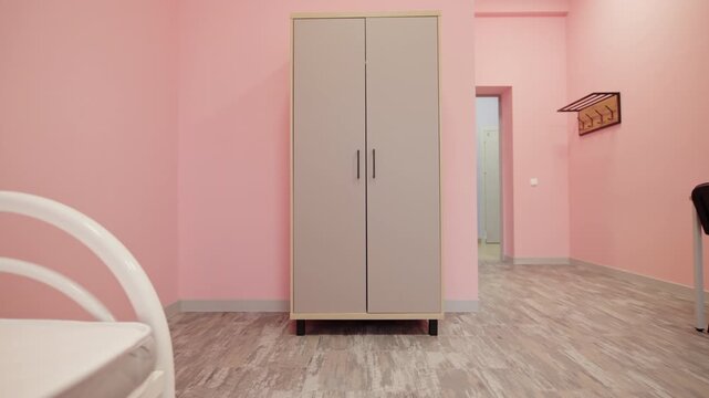 Bed frame with mattress stands in foreground of dorm room. Tall wooden wardrobe and black chairs complete minimalist room interior