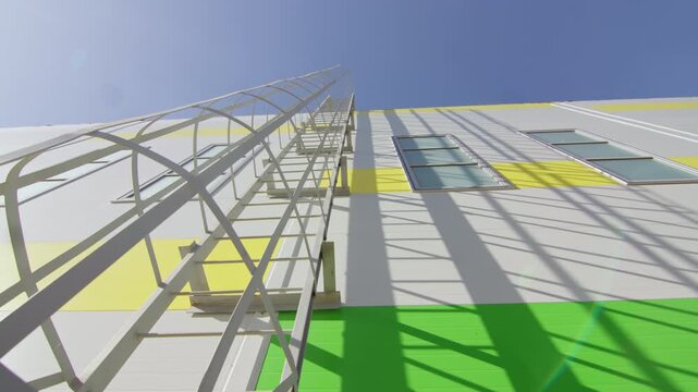 Metal safety ladder mounts on exterior wall with bright painted stripes. Strong shadows cast across building surface under bright sunny sky