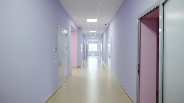 Long bright corridor features light purple walls and polished floor. Row of doors creates organized passage in modern medical facility