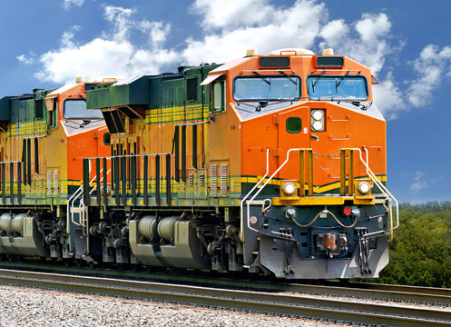 Two orange and black diesel‑electric train locomotives traveling through a green wooded corridor on a rural railroad line