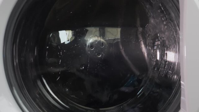 Colored items are washed in a washing machine, the drum with water and detergent is rotating. Medium shot.