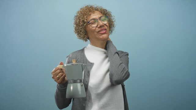 Woman holding coffeemaker, hand to neck, smiling in studio with gray cardigan and turtleneck; comfort contentment.