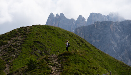 Fototapeta premium peaceful hiker standing on hillside in mountainous landscape with rocky terrain and grassy pathway, serene nature scene with green and brown hues, side view of woman wearing white shirt.