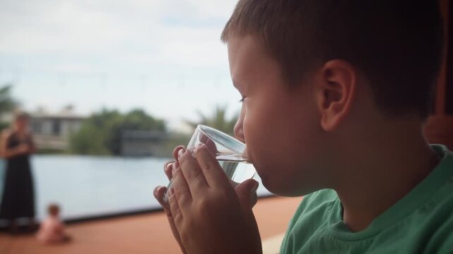 Young autistic child drinking water from a glass by a swimming pool, enjoying a calm outdoor moment with family during a summer day.