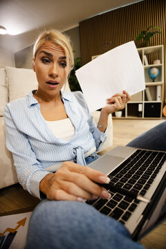 Woman Working in Home Office While Looking Surprised at Laptop Screen and Holding Paper in Small Workspace