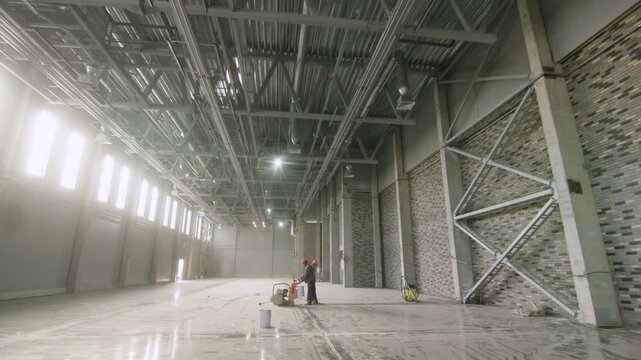 Steel support beams and brick wall decoration define spacious industrial interior. Workers perform floor finishing operations in large hall