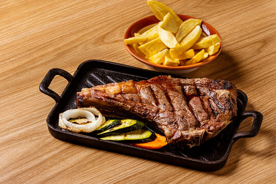 Grilled Ribeye Bone-In Steak Served on Cast Iron Skillet with French Fries, Grilled Vegetables, and Chimichurri Sauce