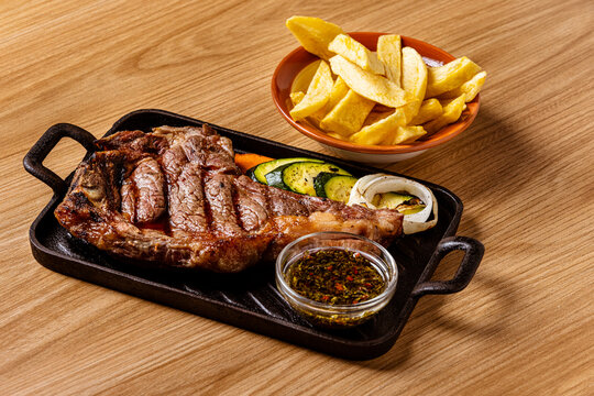 Grilled Ribeye Bone-In Steak Served on Cast Iron Skillet with French Fries, Grilled Vegetables, and Chimichurri Sauce