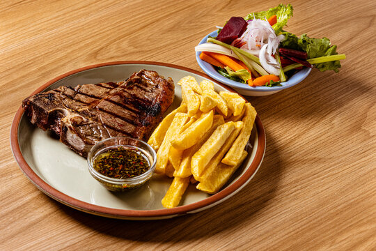 Grilled T-Bone Steak Served with Crispy French Fries and Chimichurri Herb Sauce on Ceramic Plate