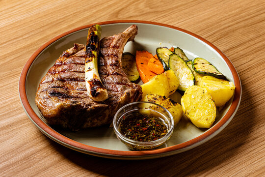 Juicy Grilled Ribeye Cowboy Steak Served with Roasted Potatoes, Grilled Zucchini, Carrots, and Chimichurri Sauce