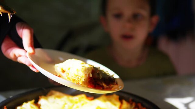 Parent serving a hot slice of homemade lasagna to a hungry expecting little boy at dinner